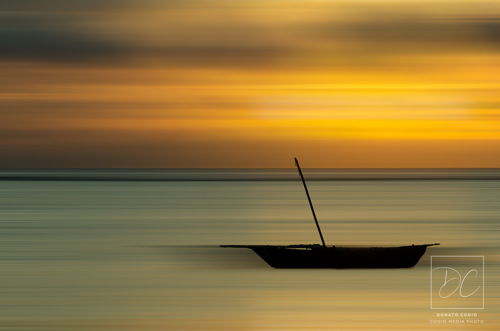 Golden Serenity – Boot im Morgenlicht | Das Bild zeigt ein ruhiges Boot vor der Küste Sansibars, umgeben von den warmen Farben des Sonnenaufgangs. Durch digitale Bearbeitung werden die Farben intensiviert, während die digital hinzugefügte Bewegungsunschärfe dem Bild eine sanfte Dynamik verleiht.Bringe die friedliche Atmosphäre eines tropischen Morgens in dein Zuhause mit meinem Fine Art Print. Dieses Kunstwerk kombiniert natürliche Schönheit mit digitaler Kunstfertigkeit und verleiht jedem Raum eine entspannende Note. - Realisiert mit Pictrs.com