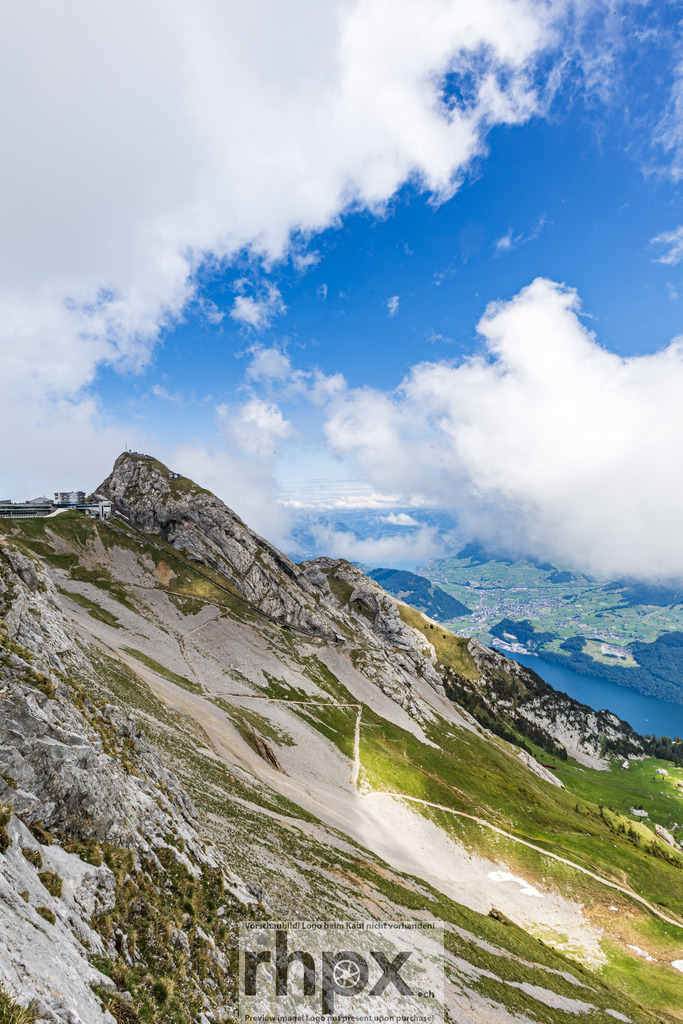 Pilatus Gipfelgrat mit Wanderweg, Vierwaldstättersee und Tal | Blick vom Grat des Pilatus auf den Hotelkomplex Pilatus-Kulm (Eselsgipfel, 2132 m ü. M.) links oben. Im Mittelgrund sichtbarer Wanderweg auf dem Grashang. Im Hintergrund Vierwaldstättersee mit Ortschaft und grünem Alpental. - Realisiert mit Pictrs.com
