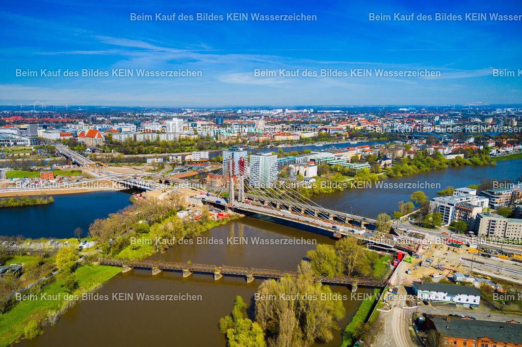 Magdeburg Pylonbrücke über Alte Elbe und Zollbrücke-0008 | Das Luftbild zeigt die Kaiser-Otto-Brücke in Magdeburg aus einem Tragschrauber. Im Vordergrund verläuft die historische Eisenbahnbrücke der ehemaligen Kanonenbahn, während sich dahinter die moderne Schrägseilkonstruktion der Kaiser-Otto-Brücke über die Alte Elbe spannt. Aus der Vogelperspektive entsteht ein eindrucksvoller Vergleich zweier Epochen des Brücken- und Verkehrsbaues.Die Kaiser-Otto-Brücke wurde zwischen 2020 und 2024 errichtet und besitzt eine Gesamtlänge von 247,99 Metern. Zwei 63 Meter hohe Pylone tragen den Brückenüberbau über insgesamt 28 Schrägseile. Die Brücke verfügt über Fahrstreifen für den Straßenverkehr, Gleise für die Straßenbahn sowie getrennte Geh- und Radwege und zählt zu den modernsten Brückenbauwerken Sachsen-Anhalts.Direkt daneben verläuft die historische Eisenbahnbrücke der ehemaligen Kanonenbahn. Die Strecke Berlin–Blankenheim wurde ab 1877 als strategische Militärbahn gebaut und diente vor allem dem schnellen Truppentransport innerhalb des Deutschen Reiches. Die genietete Stahlkonstruktion der Brücke steht bis heute als Zeugnis der Ingenieurbaukunst des späten 19. Jahrhunderts und bildet einen spannenden Kontrast zur modernen Schrägseilbrücke.Die Aufnahme aus dem Tragschrauber verbindet eindrucksvoll historische Eisenbahntechnik mit zeitgenössischem Brückenbau und dokumentiert den Wandel der Verkehrsinfrastruktur in Magdeburg über einen Zeitraum von mehr als 140 Jahren. - Realisiert mit Pictrs.com
