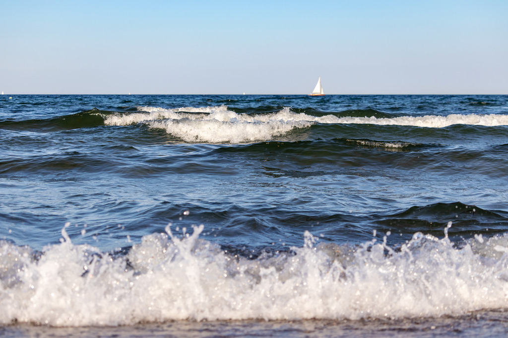 Wandbild: Wellen am Strand im Sommer | Dieses Wandbild im Querformat zeigt Wellen am sommerlichen Strand. Auf dem Meer sind einige Schaumkronen zu sehen und im Vordergrund bricht in der Unschärfe eine Welle. In der Ferne am Horizont kann man ein Segelboot sehen. - Realisiert mit Pictrs.com