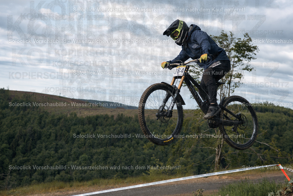 Action im Bikepark Winterberg | Ein Mountainbiker, der im Rahmen des iXS Downhill Cups 2025 im Bikepark Winterberg über eine Sprungrampe springt, wird in diesem dynamischen Foto eingefangen. Der Fahrer trägt eine komplette Schutzausrüstung und ist in der Luft, während er die Sprungrampe verlässt. Der Hintergrund zeigt eine bewaldete Landschaft mit einem bewölkten Himmel. - Realisiert mit Pictrs.com