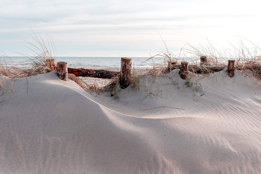Maritime Fotokunst – Helles Wandbild Sanfte Dünenlandschaft an der Ostsee | Sanfte Sandwellen schmiegen sich an verwittertes Holz, während der weite Horizont die Gedanken zur Ruhe kommen lässt. Dieses Motiv bringt die zeitlose Eleganz der Küste direkt in den Raum und schafft eine Atmosphäre von Klarheit und tiefer Entspannung. Ein Blickfang, der den hektischen Alltag vergessen lässt und die sanfte Melodie des Meeres spürbar macht.