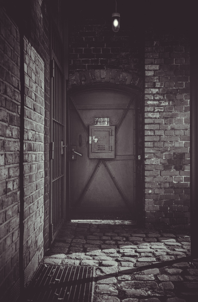 Silent Entrance  | Eine minimalistische Schwarzweißaufnahme eines verborgenen Eingangs in einem industriell geprägten Innenhof. Die raue Backsteinarchitektur, das schwere Metalltor und das einzelne, harte Licht schaffen eine dichte, beinahe filmische Atmosphäre.Licht und Schatten formen eine stille, spannungsreiche Szene, in der sich Struktur, Materialität und Raum zu einer eindringlichen Komposition verbinden. Die reduzierten Formen und die klare Linienführung lenken den Blick auf das Wesentliche und verstärken den Eindruck von Abgeschiedenheit und Geheimnis.Diese Aufnahme übersetzt urbane Architektur in eine atmosphärische Fine-Art-Komposition und eignet sich besonders für moderne, charakterstarke Räume mit einem Hang zu Kontrast und Tiefe. - Realisiert mit Pictrs.com