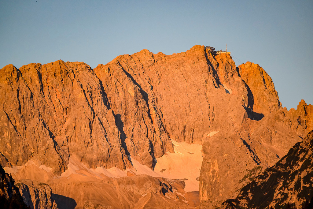 Krone der Heimat: Das goldene Leuchten der Zugspitze | Ein Anblick von überwältigender Urgewalt und tiefer, innerer Ruhe, der das Herz berührt: Im warmen, zarten Licht des Sonnenaufgangs erstrahlt das majestätische Felsmassiv der Zugspitze in einem makellosen Alpenglühen. Die schroffe Steilwand und jede feine Nuance des uralten Gesteins leuchten in sanften Gold- und Orangetönen. Hoch oben am Gipfel thront die Bergstation, hell erleuchtet im ersten Licht des erwachenden Tages, während die letzten weißen Schneefelder im Kar sanft hervortreten. Diese Aufnahme fängt die faszinierende Morgenstimmung im Hochgebirge perfekt ein – ein glanzvoller Moment voller Klarheit, Sehnsucht und Ewigkeit. - Realisiert mit Pictrs.com