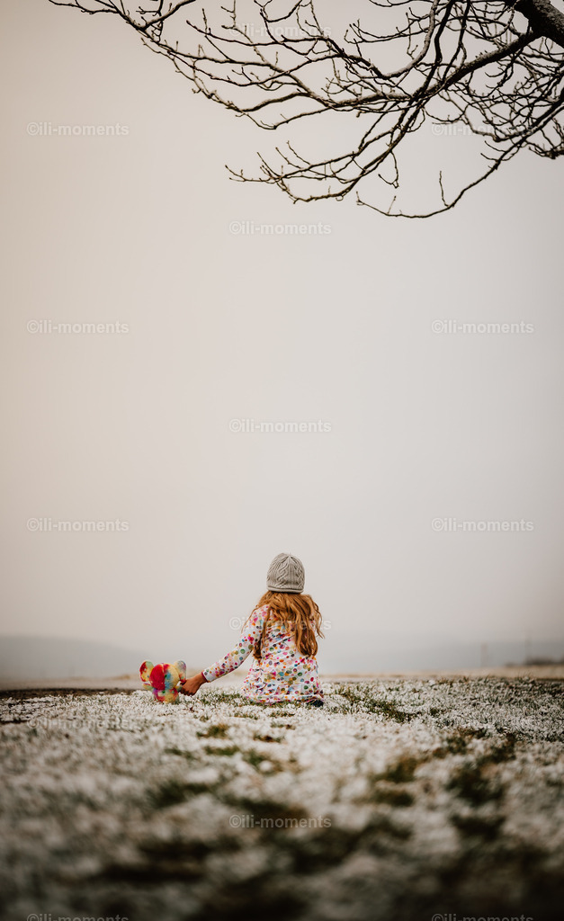 Zusammen allein | Ein Kind sitzt mit seinem Plüschtier auf einer winterlichen Wiese und blickt in die neblige Ferne. Sanfte Farbakzente verleihen dem ruhigen Bild Wärme und Nostalgie – ideal als Kunstdruck für behagliche Räume. - Realisiert mit Pictrs.com