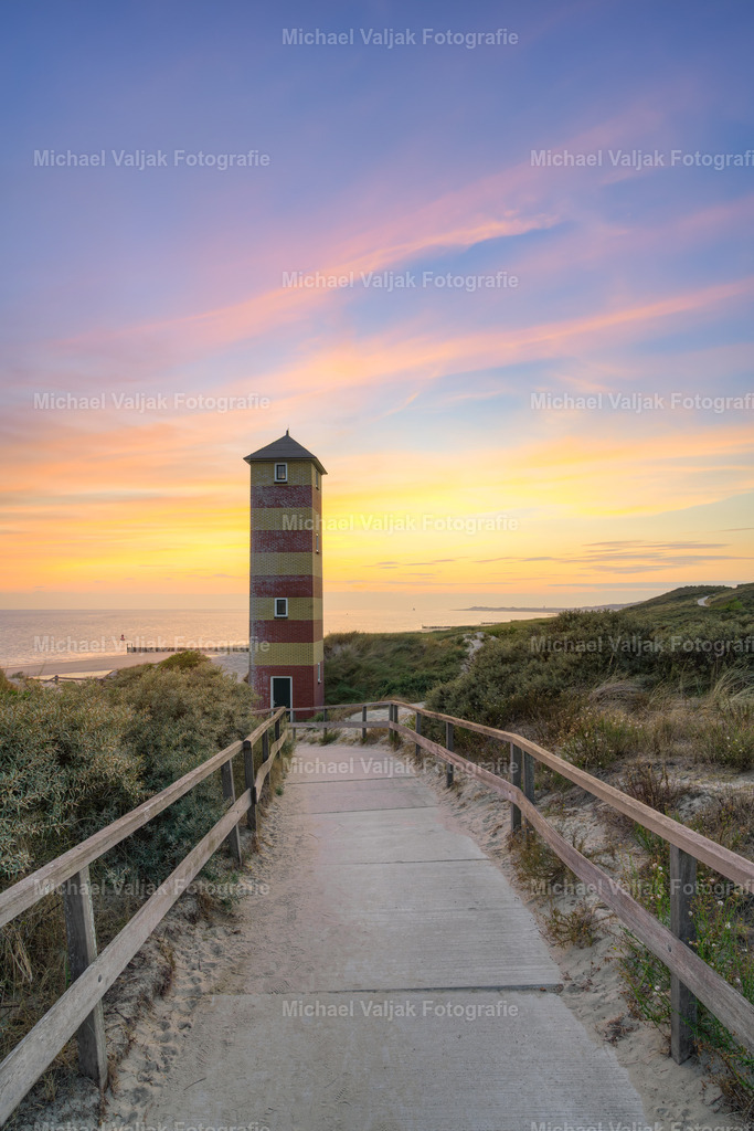 Leuchtturm Dishoek nach Sonnenuntergang | Im Hochformat führt der Weg durch die Dünen direkt auf den markanten Leuchtturm von Dishoek zu. Das Unterfeuer ragt eindrucksvoll in den noch hellen Abendhimmel und bildet den zentralen Blickpunkt der Aufnahme.Die Sonne ist bereits untergegangen, doch der Himmel leuchtet noch intensiv in Gelb, Orange und Rot. Die warmen Farben des Abendhimmels stehen dabei in schönem Kontrast zu den dunkleren Silhouetten der Dünen und dem Leuchtturm.Der nach vorne führende Weg verstärkt die räumliche Tiefe und lenkt den Blick unmittelbar auf das Leuchtfeuer. Ein stimmungsvolles Küstenmotiv aus Dishoek in Zeeland, Niederlande, das die besondere Atmosphäre der kurzen Zeit zwischen Sonnenuntergang und blauer Stunde einfängt. - Realisiert mit Pictrs.com