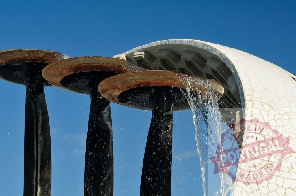 Water flowing from modern artistic fountain sculpture | Water flowing over mushroom-shaped metal elements into a white mosaic basin under a blue sky - Realisiert mit Pictrs.com