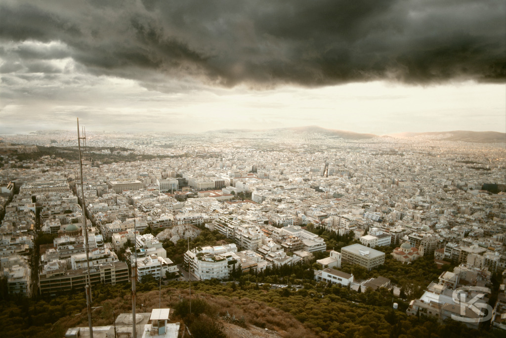 Dramatisches Luftbild von Athen unter Sturmwolken, aufgenommen vom Lycabettus Hill (2007) | Eine dramatische Luftaufnahme der dicht besiedelten Metropole Athen vom Lycabettus Hill (Lykavittos) aus, aufgenommen um das Jahr 2007. Die Aufnahme zeigt das massive, weiß getünchte Stadtbild unter einem bedrohlichen, dunkelgrauen Sturmhimmel, der einen starken Kontrast zur Helligkeit am Horizont bildet. - Realisiert mit Pictrs.com