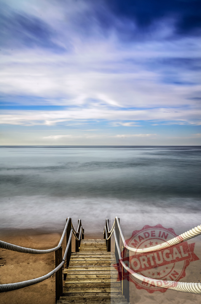 Wooden stairs leading to calm ocean waves | Wooden boardwalk steps descending to a tranquil beach and blurred ocean horizon under cloudy sky - Realized with Pictrs.com