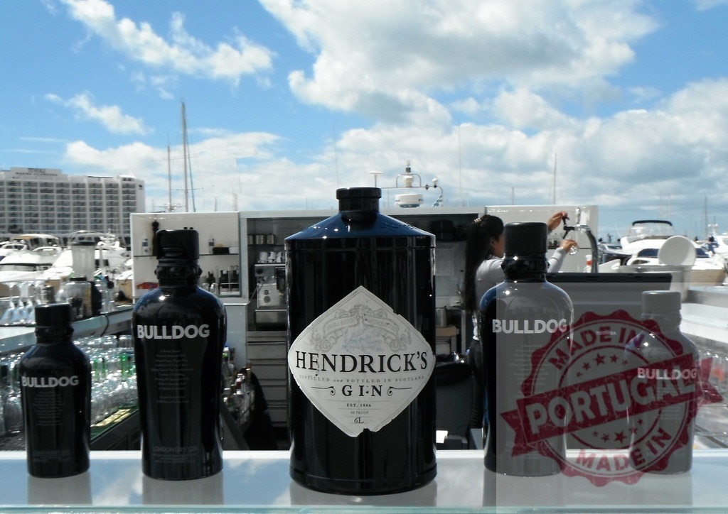 Hendrick's and Bulldog gin bottles at outdoor marina bar | Gin bottles lined up at an outdoor bar in a sunny marina, offering refreshing drinks - Realisiert mit Pictrs.com