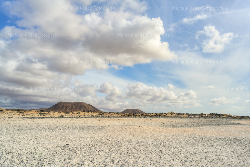 Playa del Bajo de la Burra auf Fuerteventura - Popcorn Beach | Am Playa del Bajo de la Burra, dem bekannten Popcorn Beach, liegt die Küste voller heller, rund geschliffener Algenkalkstücke, die wie verstreutes Popcorn wirken. Zwischen den weißen Formen schimmert das dunkle Lavagestein, im Hintergrund erhebt sich der Montana de la Mancha. - Realisiert mit Pictrs.com