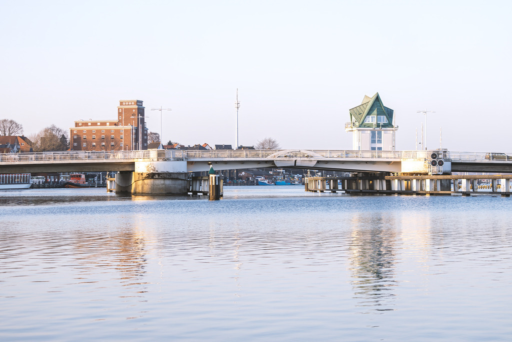 Wandbild: Norddeutsches Licht – Schleibrücke in Kappeln | Dieses Wandbild zeigt die Schleibrücke in Kappeln in einem freundlichen, hellen Licht, das die weite Wasserfläche und die klare Atmosphäre der Schlei betont. Der markante Brückenturm mit seinem grünen Dach setzt einen ruhigen architektonischen Akzent, während die umliegenden Gebäude sich harmonisch in die helle Szenerie einfügen. Das Motiv wirkt offen, leicht und typisch norddeutsch – ideal für Räume, die ein Gefühl von Weite, Klarheit und maritimer Gelassenheit ausstrahlen sollen.