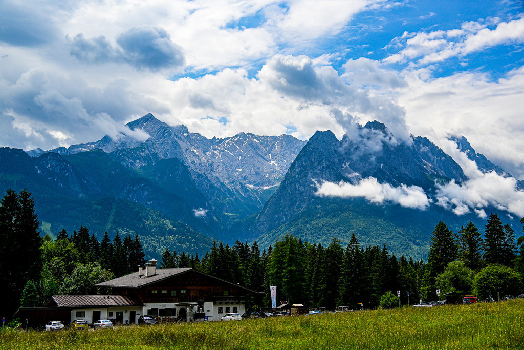 Drama am Himmel: Die ungezähmte Kraft der Berge | Ein Anblick voller Urgewalt und packender Dynamik: Majestätisch und wild erhebt sich das gewaltige Wettersteingebirge mit der Alpspitze und dem Zugspitzmassiv. Wolkenfetzen hängen wie mystische Schleier in den schroffen, felsigen Steilwänden und verleihen der Szenerie eine dramatische Tiefe. Im Kontrast dazu ruht im Vordergrund ein idyllisches alpenländisches Anwesen, eingebettet in saftig grüne Bergwiesen und dichte Nadelwälder. Diese Aufnahme fängt das faszinierende Wechselspiel zwischen friedlicher Heimatidylle und der rauen, unberechenbaren Schönheit des Hochgebirges perfekt ein. Ein Bild, das Ehrfurcht einflößt und Sehnsucht weckt. - Realisiert mit Pictrs.com