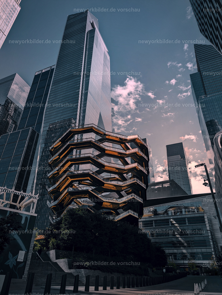 The Vessel in Hudson Yards von unten fotografiert, strahlend blauer Himmel und Sonnenlicht hinter den Gebäuden in Manhattan | Am Fuße von The Vessel erhebt sich die futuristische Struktur wie ein Kunstwerk aus Glas und Stahl. Der Himmel leuchtet in kräftigem Blau, während die Sonne hinter den Hochhäusern hervorstrahlt und goldene Reflexe auf die geschwungenen Linien wirft. Ein Moment, der die moderne Seele von New York einfängt