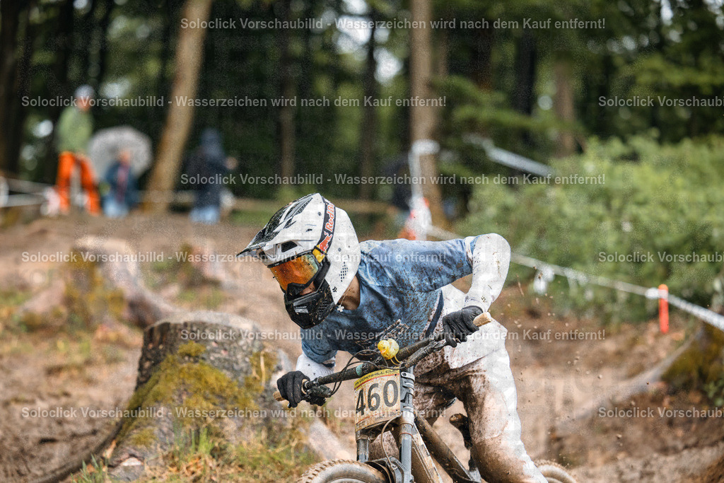 Mountainbiker im Wald | Ein Mountainbiker in voller Ausrüstung rast durch einen verschneiten Wald. Er trägt einen weißen Helm mit gelben Akzenten und eine Schutzbrille. Sein Trikot ist mit Schlamm bedeckt, und er trägt eine Nummer 460. Der Hintergrund besteht aus Bäumen und einem verschneiten Untergrund. - Realisiert mit Pictrs.com