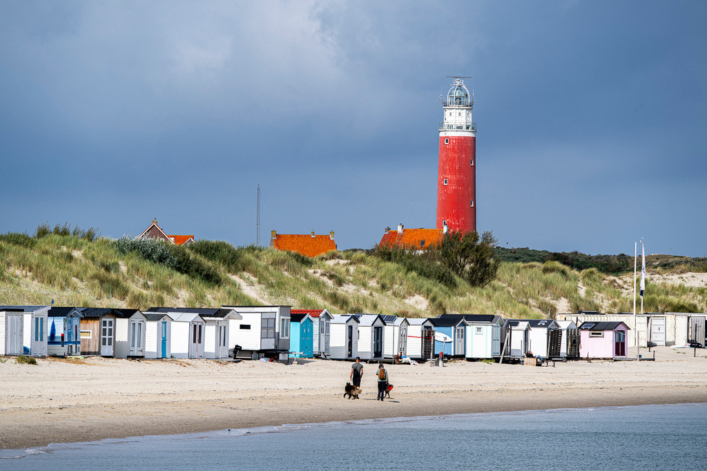 Wahrzeichen von Texel: Roter Leuchtturm und bunte Strandhäuschen an den Dünen von De Cocksdorp | Der markante rote Leuchtturm Eierland ragt hinter den begrünten Dünen am Nordseestrand von Texel auf. Entlang des Sandstrandes erstreckt sich eine Reihe farbenfroher Strandhäuschen, vor denen Spaziergänger mit Hund unterwegs sind. Ein dunkler Wolkenhimmel kontrastiert mit dem Sonnenlicht auf der Küstenlandschaft. Vielseitig einsetzbar für Berichte über Tourismus, Ausflugsziele in den Niederlanden, Inselurlaub und maritime Wahrzeichen. - Realisiert mit Pictrs.com