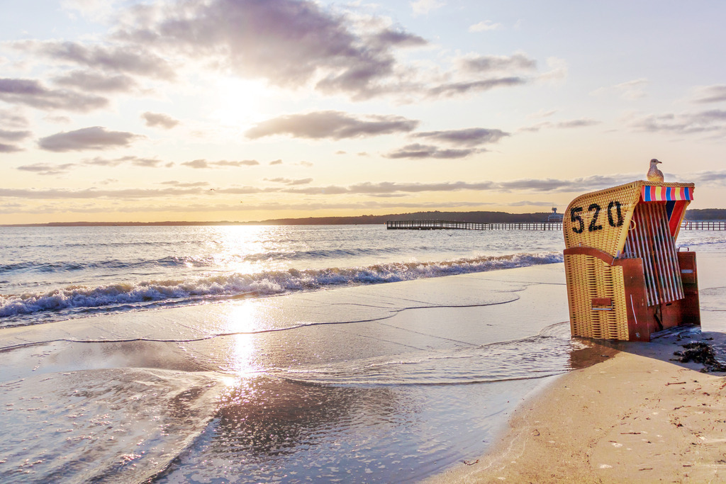 Akustikbild: Möwe auf einen Strandkörb beim Sonnenaufgang | Dieses malerische Akustikbild im Querformat zeigt einen Sonnenaufgang am Meer. Die Sonne spiegelt sich auf dem Meer und taucht den Himmel in einen schönen Verlauf aus rot bis hellblau. Am Strand steht ein Strandkorb, der durch das leichte Hochwasser teilweise im Meer steht. Auf dem Strandkorb sitzt eine Möwe.  - Realisiert mit Pictrs.com