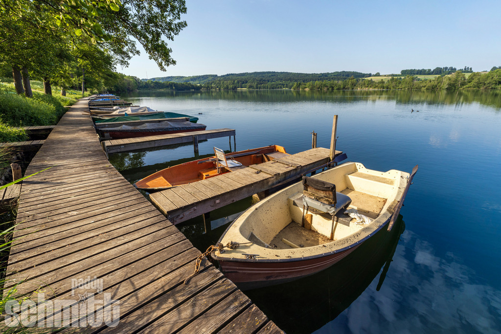 Bootssteg am Illmensee im Sommer | Ein idyllischer Bootssteg ragt am Illmensee ins ruhige Wasser und lädt an warmen Sommertagen zum Verweilen ein. Das klare Wasser, das satte Grün der Uferlandschaft und die sommerliche Stimmung vermitteln Ruhe und Erholung inmitten der Natur. - Realisiert mit Pictrs.com
