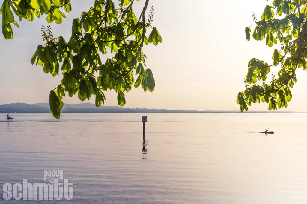Kajak auf dem Bodensee bei Lindau | Ein Kajak gleitet im warmen Abendlicht ruhig über den Bodensee bei Lindau, während die untergehende Sonne die Landschaft in goldene Farben taucht. Die stille Wasseroberfläche und die besondere Stimmung des Sonnenuntergangs vermitteln ein Gefühl von Freiheit, Ruhe und Sommerabend. - Realisiert mit Pictrs.com