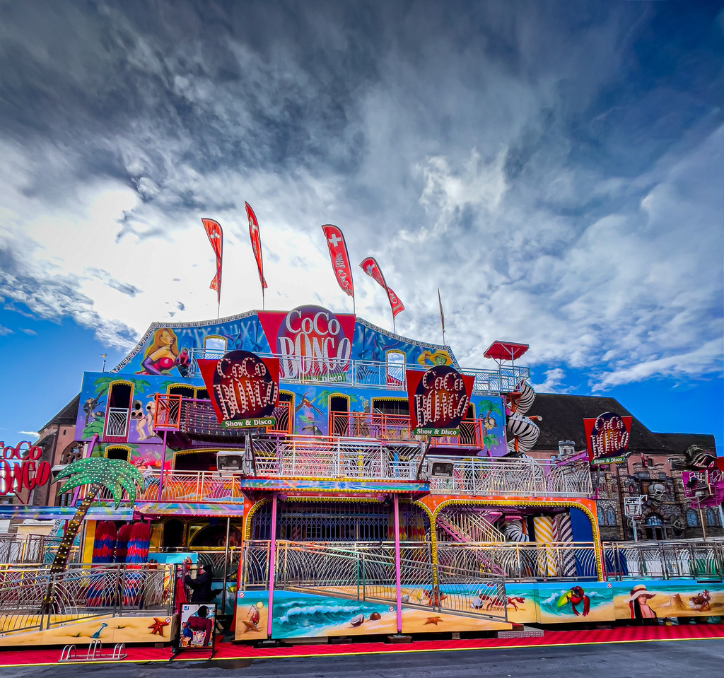 Basel Herbstmesse – Farbenfrohes Coco Bongo Laufgeschäft bei Tag | Lebendige und detailreiche Jahrmarktfotografie aus Basel: Dieses hochwertige Wandbild zeigt das kultige Laufgeschäft Coco Bongo auf der Basler Herbstmesse. Die farbenprächtige, karibische Kulisse unter strahlend blauem Wolkenhimmel bringt gute Laune und energiegeladene Dynamik in jeden Raum. Ein perfekter, bunter Blickfang als Galerie-Druck auf Leinwand oder Alu-Dibond – ideal für Kinderzimmer, moderne Hobbyräume oder als Geschenk für Basel-Liebhaber. - Realisiert mit Pictrs.com