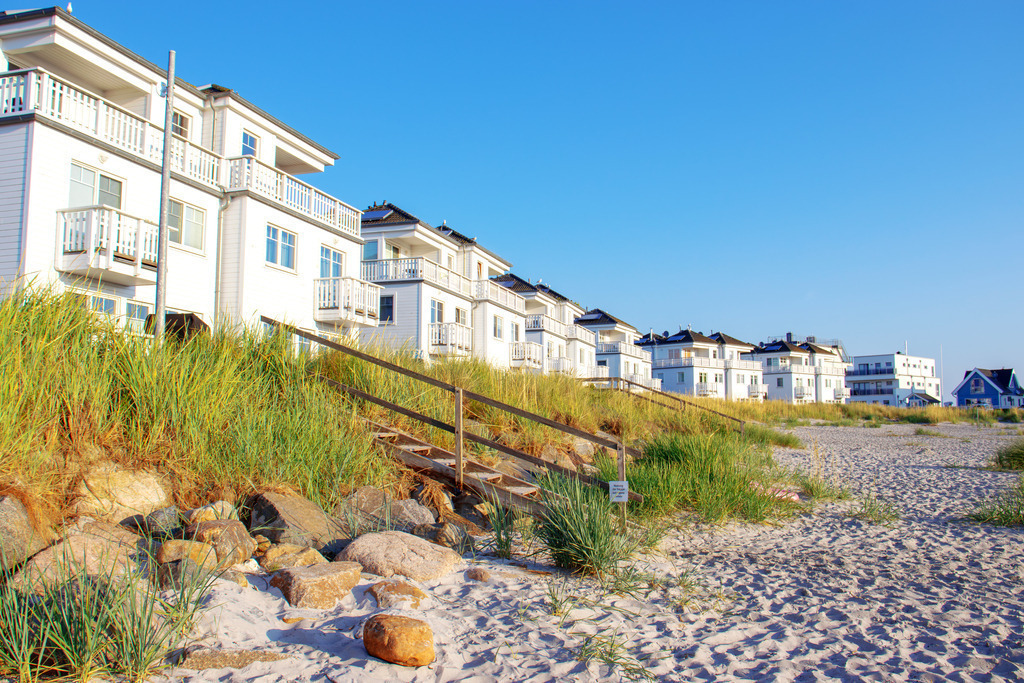Wandbild: Strand in Olpenitz im Sommer | Dieses Wandbild im Querformat zeigt den Sandstrand in Olpenitz im Sommer. Direkt am Sandstrand befinden sich Strandhafer und eine Treppe zu anliegenden Ferienwohnungen. Der blaue Himmel ist wolkenlos.  - Realisiert mit Pictrs.com