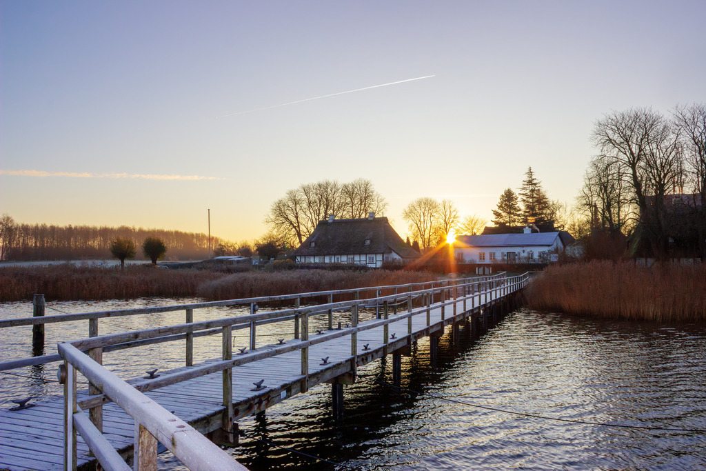 Wandbild: Sonnenaufgang an einem frostigen Wintertag in Sieseby | Dieses Wandbild im Querformat zeigt einen schönen Sonnenaufgang an einem frostigen Wintertag in Sieseby an der Schlei. Im Vordergrund ist der leicht vereiste Schleianleger zu sehen. Die Sonne geht zwischen den beiden malerischen Häusern am Schleiufer auf. Auch Sonnenstrahlen sind gut zu erkennen. Am Horizont ist der Himmel durch die morgendliche Lichtstimmung leicht rötlich. Der Himmel ist fast wolkenlos.  - Realisiert mit Pictrs.com