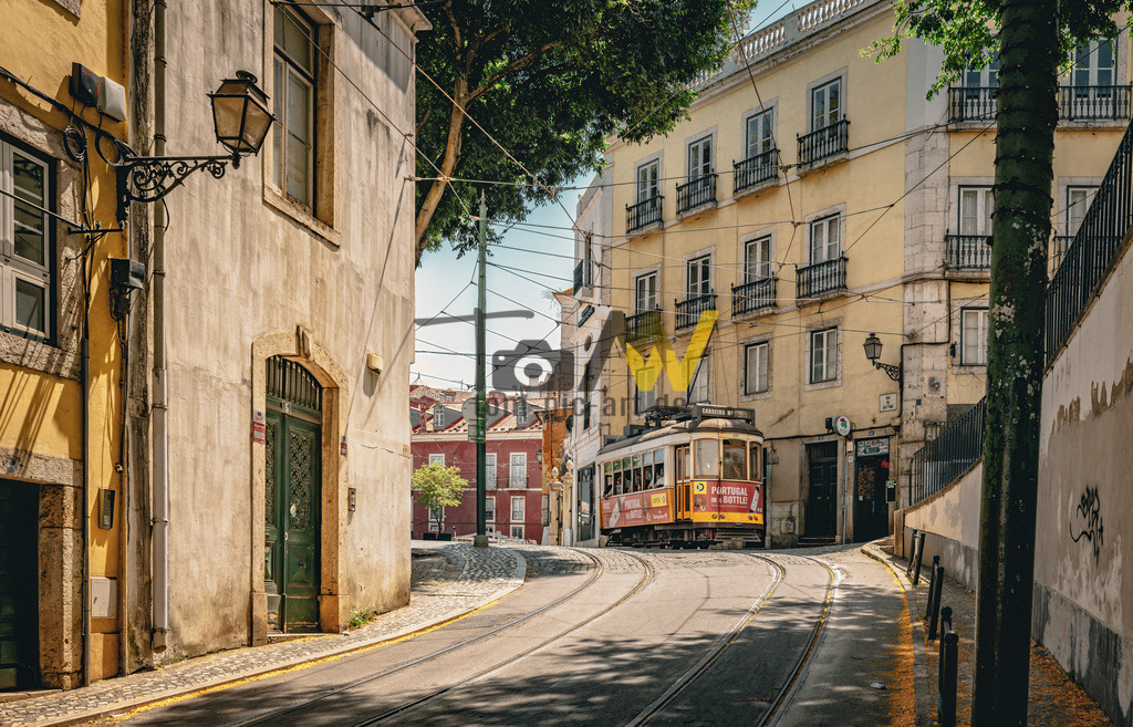 Eine "Elétrico" oder Staßenbahn fährt durch die Altsadt Lissabons | Das Bild zeigt eine Straßenbahnszene in Lissabon, Portugal.Die Szene zeigt eine ikonische gelbe Lissabonner Straßenbahn, bekannt als "Elétrico", die durch die engen, gepflasterten Straßen der Stadt fährt. Die Architektur ist typisch für die historische Baixa Pombalina oder die umliegenden Viertel Alfama/Graça. Die Straßenbahnen sind ein wesentliches Merkmal der Stadt und eine beliebte Touristenattraktion - Realisiert mit Pictrs.com