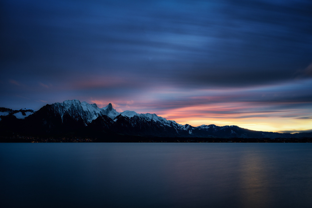 Pastell am Horizont | Eine weite Landschaftsaufnahme bei Dämmerung, dominiert von einem tiefblauen See im Vordergrund, dessen Oberfläche glatt und spiegelnd erscheint. Über dem See erstreckt sich eine schmale Reihe von funkelnden Lichtern einer Ufersiedlung. Dahinter erhebt sich eine dunkle, schneebedeckte Bergkette mit scharfen, gezackten Gipfeln, deren Flanken teilweise weiss leuchten. Der Himmel darüber ist ein Spektakel aus langgezogenen Wolkenstreifen, die von tiefem Violett und Dunkelblau am oberen Rand zu leuchtenden Rosa-, Orange- und Gelbtönen am Horizont übergehen. Die Farben des Himmels sind weich ineinander verlaufend, was auf eine Langzeitbelichtung hinweist. - Realisiert mit Pictrs.com