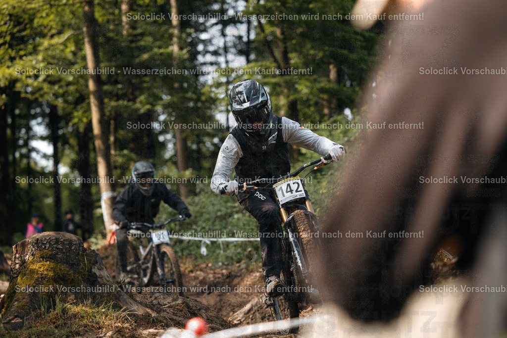 Mountainbike-Action im Wald | Ein Mountainbiker in voller Ausrüstung rast durch einen bewaldeten Kurs. Er trägt einen schwarzen Helm mit Visier und eine schwarze Weste über einem schwarzen T-Shirt. Er trägt auch schwarze Handschuhe und schwarze Knieschützer. Er trägt die Nummer 142 auf seinem Rücken. Der Hintergrund besteht aus dichtem Grün und Bäumen. Ein weiterer Mountainbiker ist im Hintergrund zu sehen. - Realisiert mit Pictrs.com