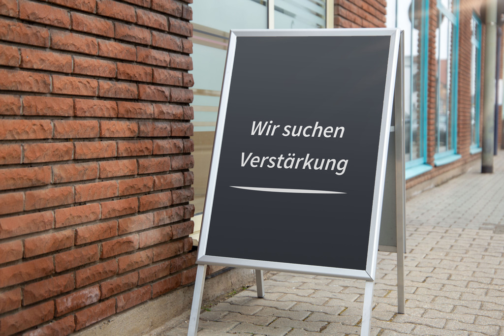 WIR SUCHEN VERSTÄRKUNG | (Fotomontage) Aufsteller mit der Aufschrift WIR SUCHEN VERSTÄRKUNG vor einem Geschäft - Realisiert mit Pictrs.com