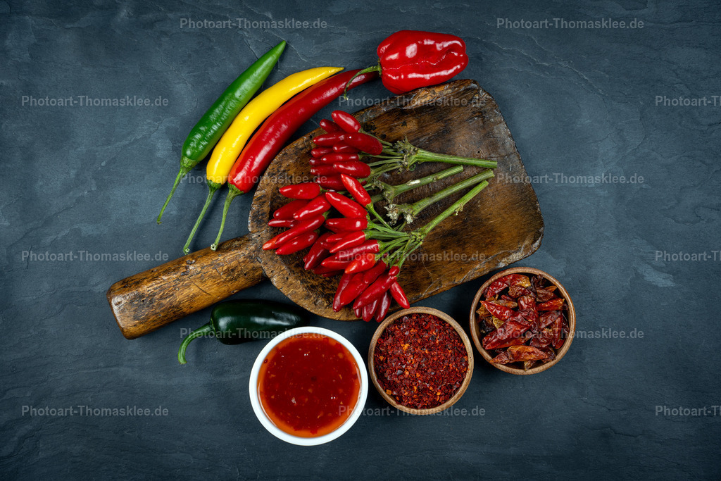 Scharfe Chili-Vielfalt auf rustikaler Holzschaufel | Eine rustikale Holzschaufel präsentiert verschiedene Chilisorten auf dunklem Schiefergrund. Frische grüne, gelbe und rote Chilischoten liegen neben getrockneten roten Exemplaren. Drei kleine Holzschalen enthalten leuchtend rotes Chilipulver und getrocknete Chiliflocken. Eine weiße Schale zeigt scharfe rote Sauce. Die warmen Rottöne der Gewürze kontrastieren wirkungsvoll mit dem kühlen Schieferuntergrund. Diese Komposition zeigt die Vielfalt scharfer Gewürze in natürlicher, appetitlicher Präsentation für Kochbegeisterte. - Realisiert mit Pictrs.com