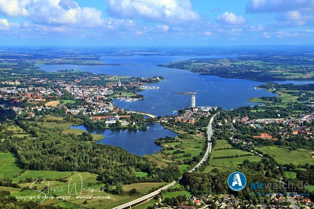 "Aerial view of Schleswig approaching from the west: A study in geographic mapping and urban development" | Luftbilder zeigen die Schlei als schmale, gewundene Wasserstraße, die die Stadt durchzieht, sowie markante Sehenswürdigkeiten wie die Schlossinsel mit dem Schleswig-Holstein-Museum, die St.-Petri-Dom und die umliegenden Wohngebiete.
