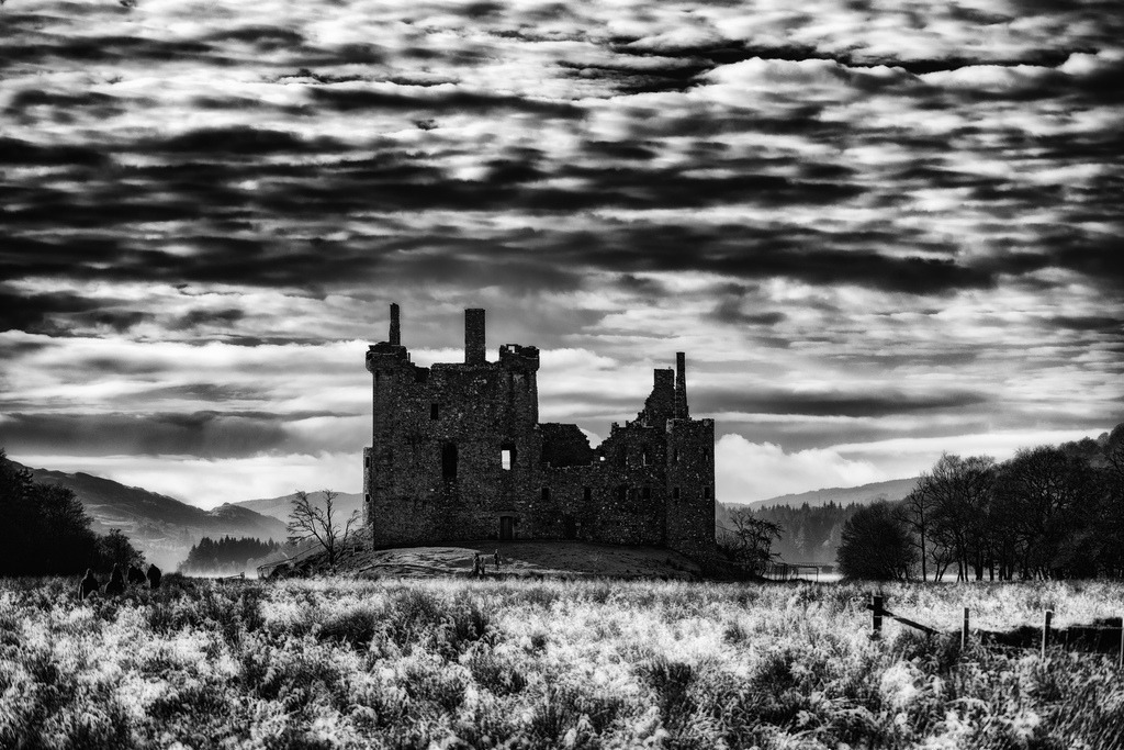 Kilchurn Castle | Die Ruine von Kilchurn Castle dominiert die winterliche Landschaft Schottlands. Eine dramatische Wolkenformation erstreckt sich über den Horizont, während der Vordergrund von gefrorenem Gras bedeckt ist. Die Schwarz-Weiss-Darstellung betont die raue Schönheit und historische Präsenz des Ortes. - Realisiert mit Pictrs.com