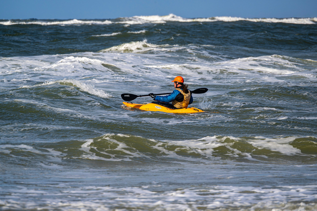Einsam | Das Bild zeigt einen Seekajakfahrer in einem gelben Boot, der sich durch die unruhige Brandung der Nordsee kämpft. Die Person trägt eine blaue Paddeljacke, eine Schwimmweste und eine leuchtend orangefarbene Kopfbedeckung, während sie das Doppelpaddel kraftvoll einsetzt. Das Meer ist von hohen Wellen und weißer Gischt geprägt, was die Dynamik und die sportliche Herausforderung des Wellenreitens unterstreicht. Der Horizont ist tief angesetzt, wodurch der Fokus ganz auf der Interaktion zwischen Mensch und Elementen liegt. - Realisiert mit Pictrs.com