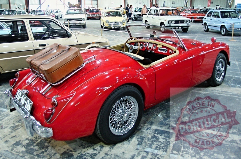 Red MG A roadster parked, showing its spoked wheels, with luggage on the trunk | Classic red MG A roadster parked in exhibition hall with luggage on the trunk rack, showing its spoked wheels - Realized with Pictrs.com