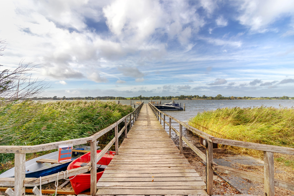 XXL Wandbild: Bootssteg am Ostseefjord | Dieses Wandbild im Querformat zeigt einen malerischen Bootssteg am Ostseefjord. Auf der linken Seite sind Boote zu sehen. Am blauen Himmel sind schöne Wolken zu sehen.  - Realisiert mit Pictrs.com