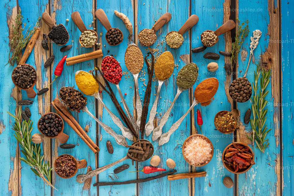 Gewürzvielfalt auf rustikalem Holz - Kulinarische Kunst II | Auf einem verwitterten blauen Holzuntergrund präsentiert sich eine kunstvolle Anordnung verschiedener Gewürze und Kräuter. Silberne Löffel und hölzerne Schaufeln tragen bunte Gewürze wie Kurkuma, Paprika, Pfeffer und getrocknete Chilis. Frische Rosmarinzweige rahmen die Komposition ein, während Zimt-Stangen und rosa Salz zusätzliche Farbakzente setzen. Diese harmonische Darstellung zeigt die Vielfalt der Küchenschätze und lädt zum Kochen ein. Die rustikale Optik verleiht dem Motiv einen authentischen Charme. - Realisiert mit Pictrs.com
