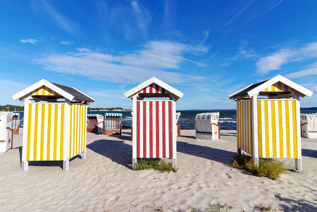 Leinwand: Farben des Sommers – Umkleidekabinen am Strand | Dieses Wandbild zeigt die farbenfrohen Umkleidekabinen am Sandstrand von Eckernförde an der Ostsee. Die Kabinen stehen in klarer Reihe, ihre vertikalen Streifen in Gelb-Weiß und Rot-Weiß leuchten im Sonnenlicht und setzen lebendige Akzente vor dem blauen Himmel. Dahinter reihen sich klassische weiße Strandkörbe entlang des Strandes – nummeriert und ausgerichtet zum Meer. Sie verleihen der Szene eine typische norddeutsche Strandatmosphäre: geordnet, ruhig und einladend. Die Komposition lebt vom Zusammenspiel aus Farbe, Struktur und Küstenkultur – ein Bild, das Sommerstimmung und maritimes Flair vermittelt. Ideal als Wandbild für helle, naturnahe Wohnkonzepte. Ein frischer, fröhlicher Akzent für Wohnzimmer, Büro oder Ferienwohnung. - Realisiert mit Pictrs.com