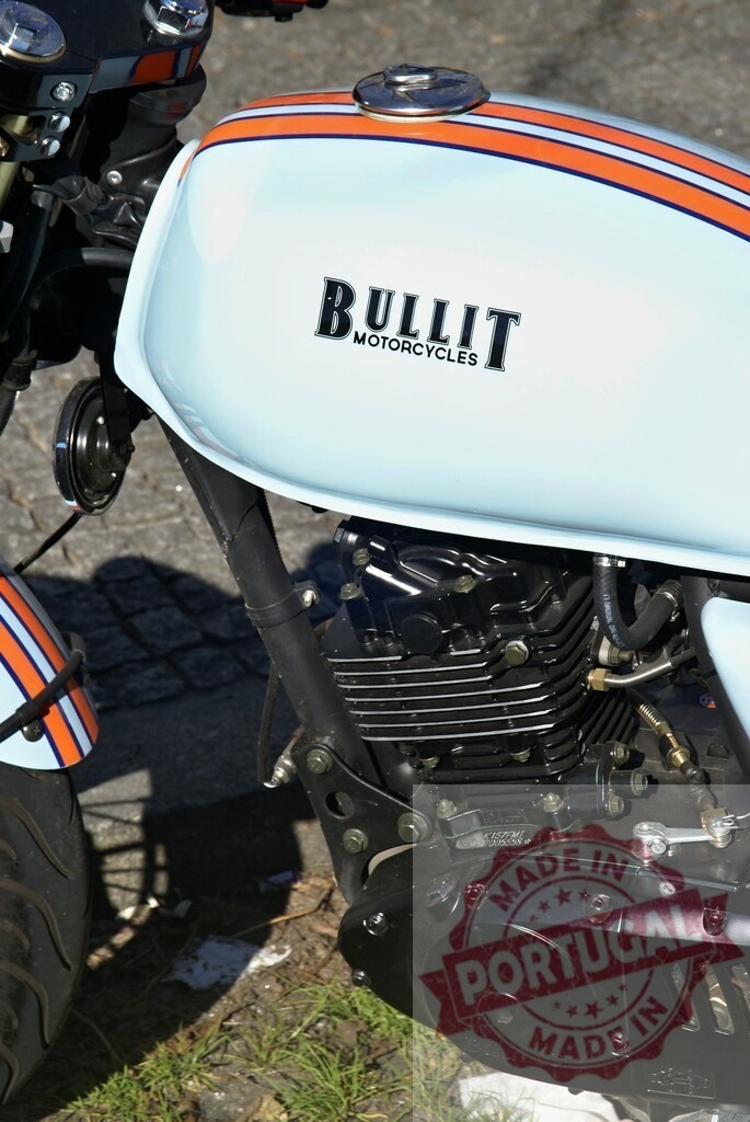 Bullit motorcycle parked on a paved road with grass on the side | Close-up of a Bullit motorcycle engine and fuel tank, parked on a paved road with grass on the side - Realized with Pictrs.com