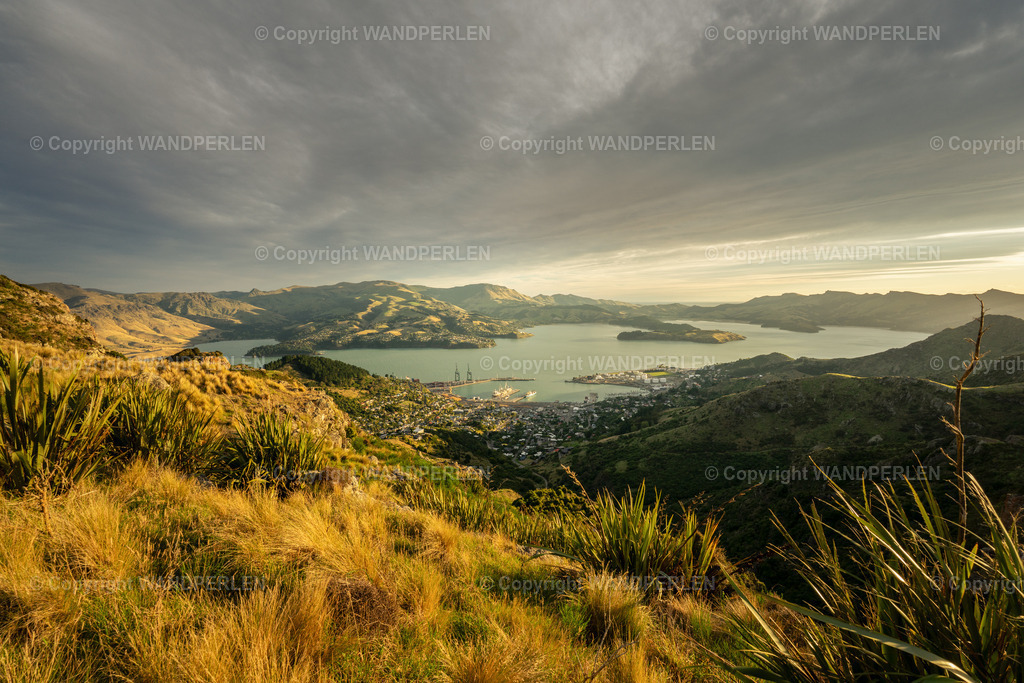 Abendsonne | Vom Berg hinunter öffnet sich der Blick auf Lyttelton und das weite Wasser des Hafens. Warmes Abendlicht überzieht die Hügel und lässt das Meer in sanften, goldenen Tönen schimmern. Darüber türmt sich ein dramatischer Wolkenhimmel in kühlen Blau- und Grautönen, der dem Motiv spannungsvolle Tiefe verleiht.Genau dieser Kontrast zwischen glühender Erde und kühler Himmelsweite macht das Bild so eindrucksvoll. - Realisiert mit Pictrs.com