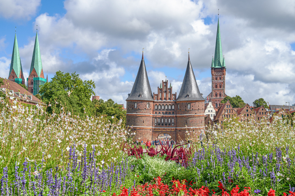 Holstentor Lübeck | Das Holstentor in Lübeck ist ein über Deutschland hinaus bekanntes Wahrzeichen dieser Stadt. Es wurde im Jahr 1478 fertiggestellt und beherbergt heute das Stadtgeschichtliche Museum von Lübeck. Die Aufnahme entstand im Herbst, Blumen und ein schöner Wolkenhimmel rahmen das historische Bauwerk romantisch ein. — Auflösung des Originals: 6015 x 4010 px. - Realisiert mit Pictrs.com