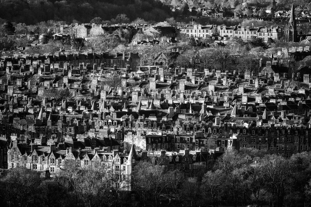Dächerlandschaft Edinburgh | Eine dichte Ansammlung historischer Gebäude und Schornsteine prägt die Hügellandschaft von Edinburgh. Die Schwarz-Weiss-Aufnahme betont die Texturen der Fassaden und Dächer sowie das Spiel von Licht und Schatten. Die Telekompression verdichtet die architektonischen Elemente zu einem komplexen Muster. - Realisiert mit Pictrs.com