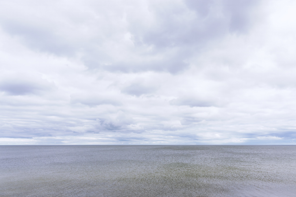 Wandbild: Ablandiger Wind über stillem Meer | Feine Kräuselungen auf der Wasseroberfläche verraten den ablandigen Wind, der sanft über das ruhige Meer streicht. Die klare Horizontlinie trennt das stille Wasser von einem wolkenreichen Himmel, dessen weiche Lichtstimmung dem Motiv eine meditative Tiefe verleiht. Die reduzierte Komposition und die subtilen Strukturen schaffen eine zeitlose, minimalistische Ästhetik, die Ruhe und Weite eindrucksvoll spürbar macht.