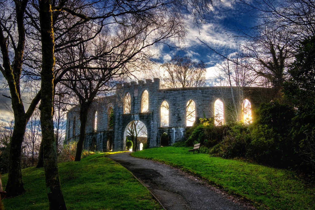 McCaig's Tower Oban | Die imposante Rundmauer von McCaig's Tower in Oban, Schottland, wird von der tief stehenden Wintersonne durchleuchtet. Ein gewundener Pfad führt durch die von kahlen Bäumen gesäumte Landschaft auf das historische Bauwerk zu. Der dramatische Himmel mit Wolken bildet einen starken Kontrast zum warmen Licht. - Realisiert mit Pictrs.com