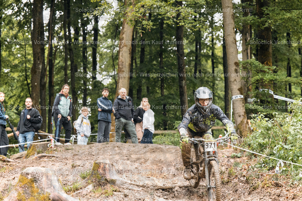 Mountainbike-Action im Wald | Ein Mountainbiker mit Helm und Schutzbrille rast auf seinem Fahrrad durch einen verschlungenen Waldpfad. Er trägt eine Nummer 371 auf seinem Fahrradrahmen. Am Rand des Pfades stehen mehrere Zuschauer, die ihn anfeuern. Der Wald ist dicht bewachsen mit Bäumen und Sträuchern. Das Licht ist gedämpft und erzeugt eine dramatische Atmosphäre. - Realisiert mit Pictrs.com
