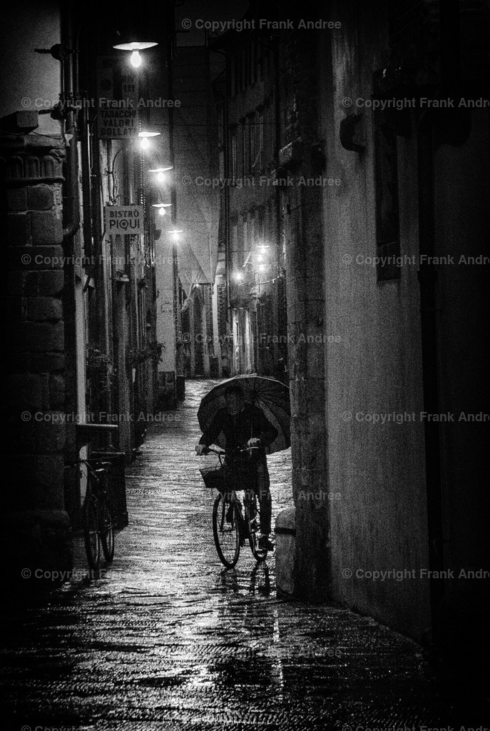 Regen in Lucca | Ein regnerischer Abend in der beleuchteten Altstadt von Lucca in der Toskana. Ein Mann fährt mit einem Regenschirm in der Hand auf seinem Fahrrad durch eine enge Gasse. Straßenfotografie aus Italien in schwarz und weiß. - Realisiert mit Pictrs.com