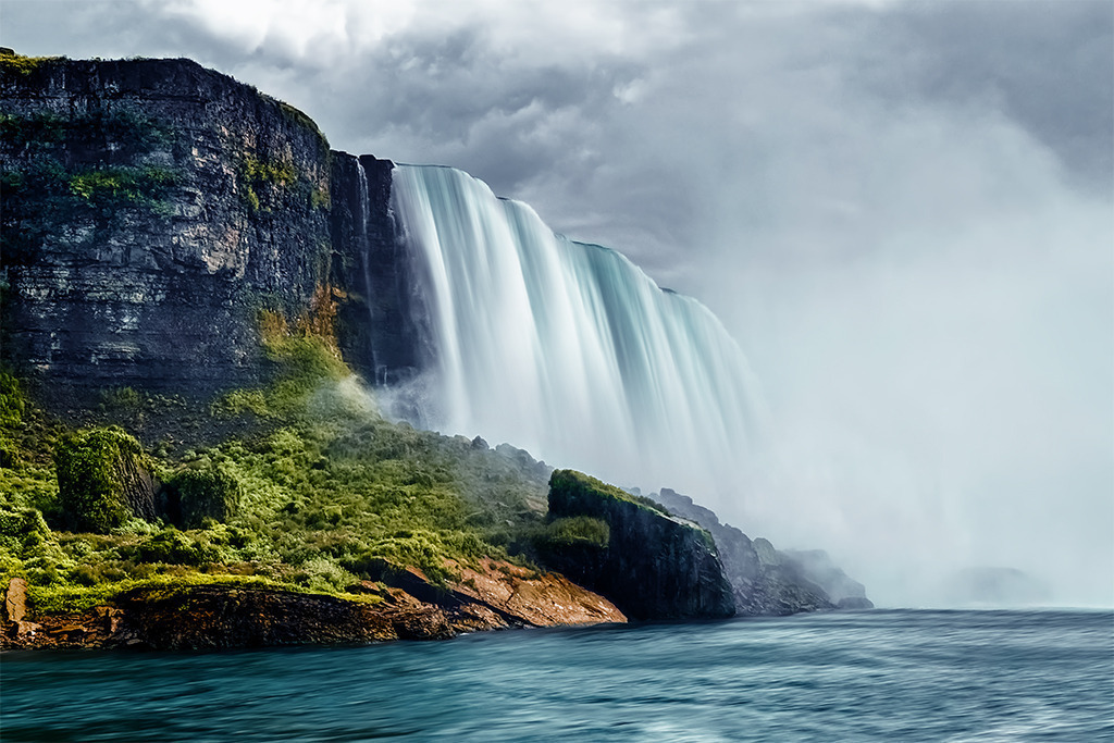 Niagarafälle, Kanada | Dichte Gischt umhüllt die kraftvollen Niagarafälle, die unter einem mit schweren Wolken verhangenen Himmel tosend in die Tiefe stürzen, während einige Sonnenstrahlen sich ihren Weg auf die beeindruckende Landschaft bahnen und die Szenerie zum Leuchten bringen.Unsere Empfehlung: Alu-Dibond oder Acrylglas - Realisiert mit Pictrs.com