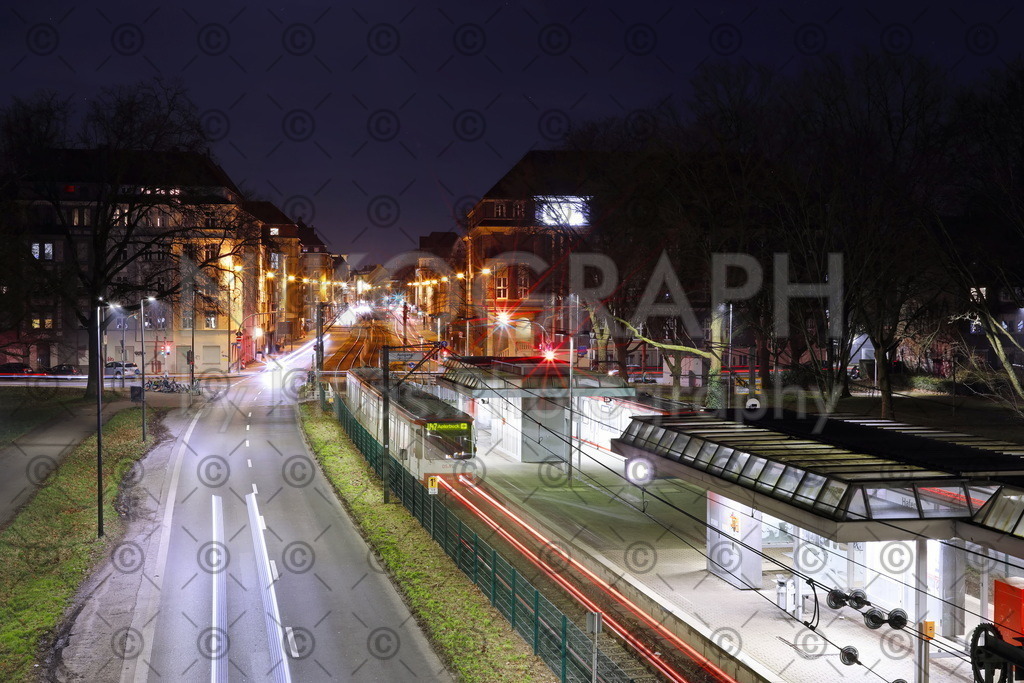 Lichtspuren in Dortmund | Auf dem Bild ist eine belebte städtische Szene aus Dortmund zu sehen, die von einer Vielzahl von Lichtspuren geprägt ist. Im Vordergrund befindet sich eine mehrspurige Strasse, auf der sich Fahrzeuge in beide Richtungen bewegen. Die Strasse ist gut beleuchtet, und die Lichter der Fahrzeuge bilden lange Spuren, die ihre Bewegungen durch die Stadt verfolgen.
Entlang der Strasse erstreckt sich eine Bahnhaltestelle für die Stadtbahn. Eine Strassenbahn ist gerade dabei, an der Haltestelle einzufahren oder abzufahren, während Passagiere ein- oder aussteigen. Die charakteristische Form der Strassenbahn ist deutlich zu erkennen, ebenso wie ihre typische Farbgebung.
Im Hintergrund erstreckt sich das städtische Panorama mit verschiedenen Gebäuden und Lichtern, die die Skyline der Stadt erhellen. Die Szene vermittelt ein Gefühl von Dynamik und Aktivität, typisch für das urbane Leben, das sich um den öffentlichen Nahverkehr und die geschäftige Strassenaktivität dreht.