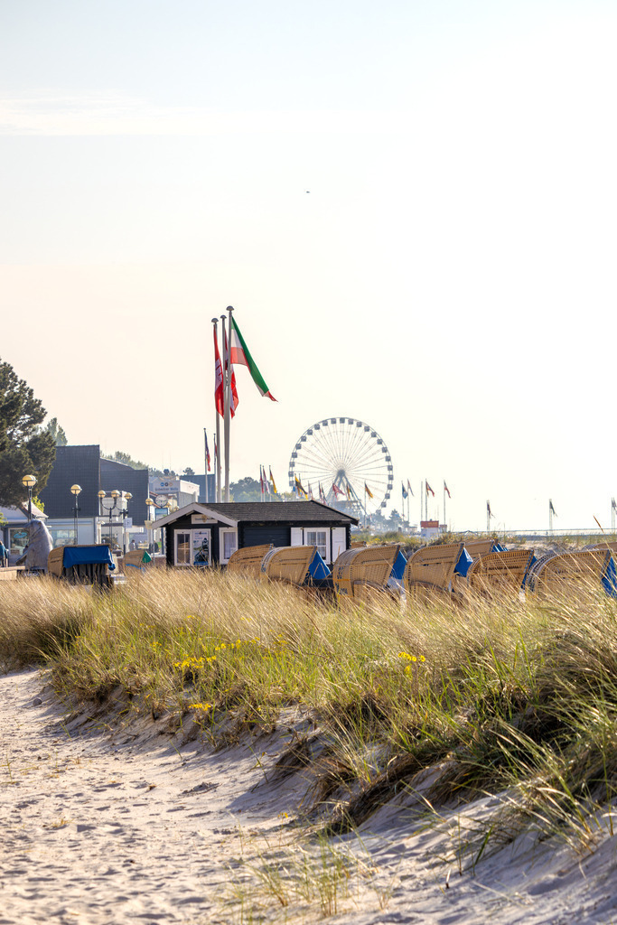 Wandbild: Zwischen Dünen und Attraktionen – Grömitzer Küstenblick | Dieses Wandbild zeigt eine lebendige Szene vom Strand in Grömitz. Ein sandiger Weg führt durch hohes Strandgras direkt zu einem belebten Strandabschnitt mit Strandkörben und kleinen Hütten. Im Hintergrund erhebt sich ein großes Riesenrad – ein markantes Wahrzeichen, das dem Bild eine festliche und familienfreundliche Atmosphäre verleiht. Mehrere Flaggen wehen im Wind und unterstreichen die internationale Offenheit des Ortes. Die Kombination aus natürlicher Vegetation, maritimer Infrastruktur und Freizeitangeboten macht dieses Motiv besonders vielseitig. Der klare Himmel und das ruhige Meer runden die Szene ab und vermitteln sommerliche Leichtigkeit. Dieses Motiv eignet sich ideal als Wandbild für maritime Wohnkonzepte – ob als Leinwandbild, Acrylglasbild, Alu-Dibond FineArt Print oder als Akustikbild. Ein stilvoller Akzent für Wohnzimmer, Büro oder Ferienwohnung. - Realisiert mit Pictrs.com