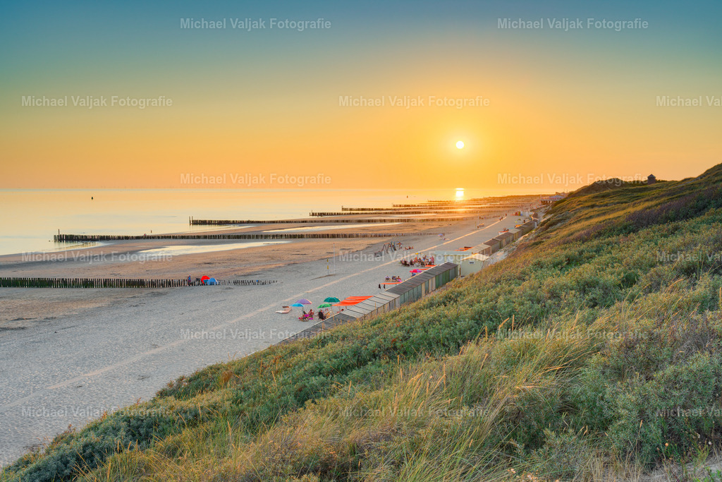 Blick von der Aussichtsdüne in Zoutelande | Von der Aussichtsdüne in Zoutelande eröffnet sich ein weiter Blick über die zeeländische Küstenlandschaft und die Nordsee. Die tief stehende Sonne taucht den Strand, die Dünen und das Meer in warmes, goldenes Abendlicht und verleiht der Szenerie eine besonders stimmungsvolle Atmosphäre.Der erhöhte Blick von der Düne macht die Weite der Küste eindrucksvoll erlebbar. Der Strand zieht sich entlang des Horizonts, während das sanfte Licht des Sonnenuntergangs die Landschaft in warme Farbtöne hüllt.Ein atmosphärisches Landschaftsfoto von Zoutelande in Zeeland, Niederlande, das die besondere Schönheit der niederländischen Nordseeküste im letzten Licht des Tages einfängt. - Realisiert mit Pictrs.com